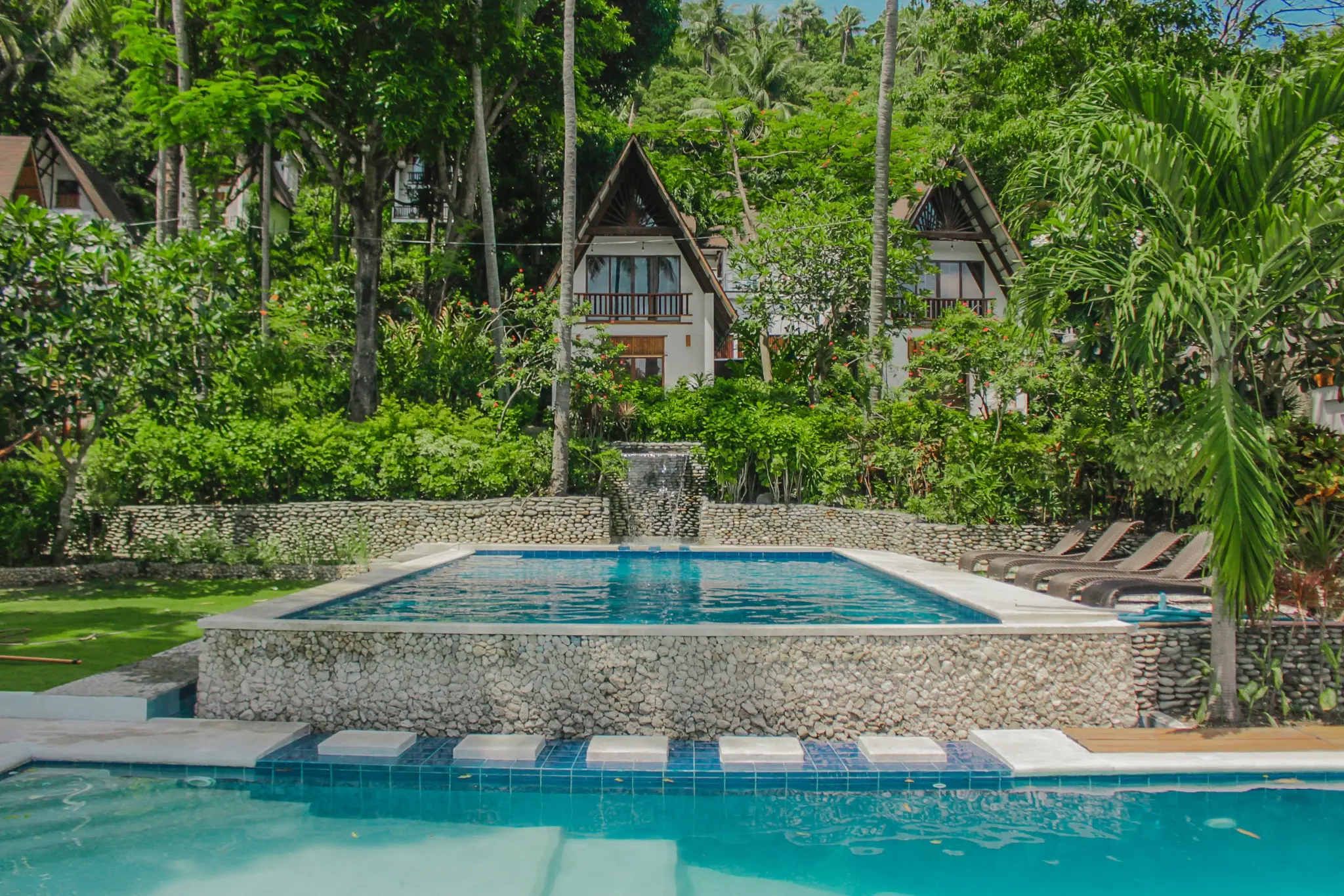 Infinity pool with cascading waterfall surrounded by lush greenery and A-frame villas at Casalay Boutique Villas & Dive Resort in Puerto Galera, Philippines.