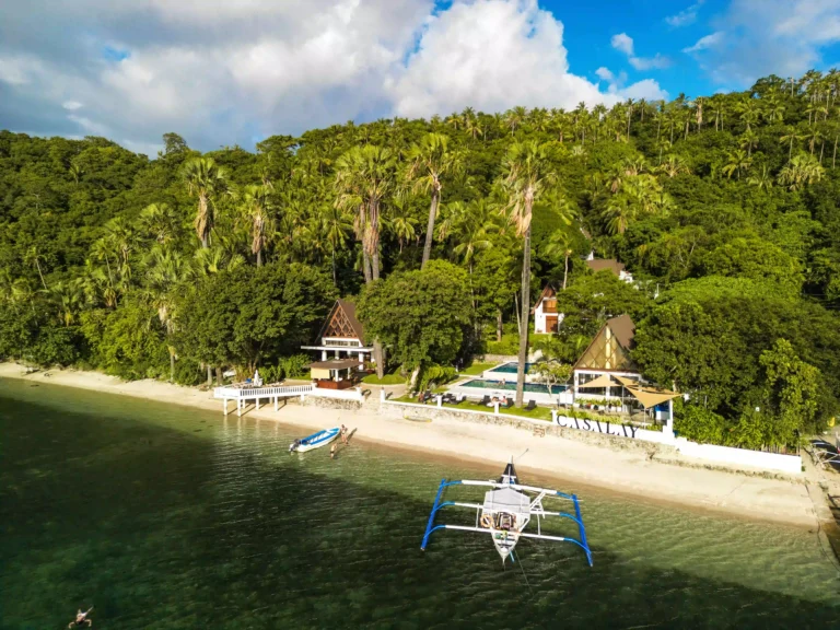Aerial view of Casalay Boutique Villas & Dive Resort in Puerto Galera, Philippines, nestled between lush green hills and a pristine white sand beach with traditional Filipino outrigger boats docked nearby.